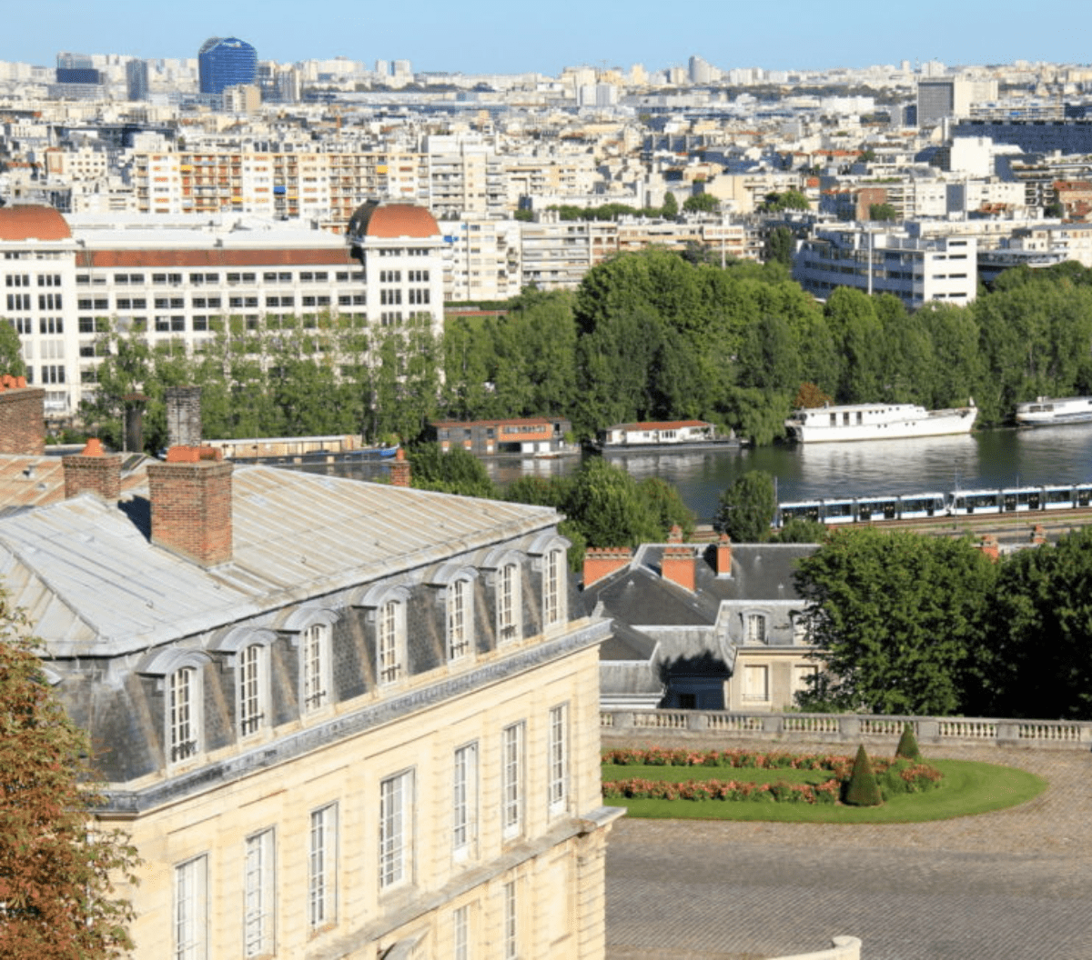 Un bâtiment historique avec une façade en pierre surplombe une rivière bordée d'arbres, avec des bateaux amarrés le long de l'eau et un paysage urbain de bâtiments modernes à l'arrière-plan.