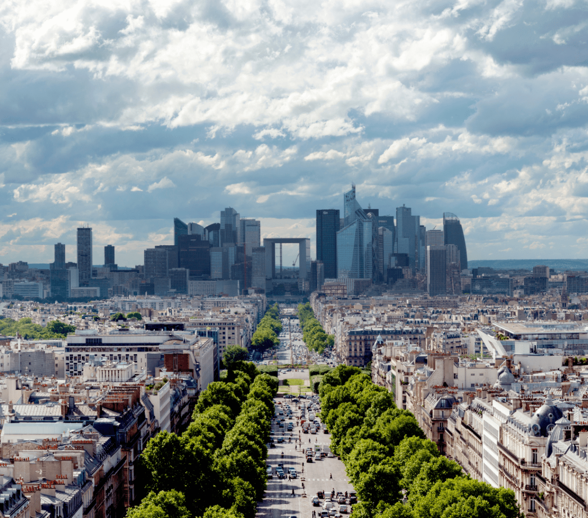 Vue de Paris avec des rues bordées d'arbres menant aux gratte-ciel modernes du quartier d'affaires de La Défense, sous un ciel partiellement nuageux.