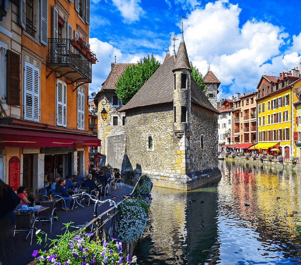 Un bâtiment médiéval en pierre se trouve au bord d'un canal entouré de bâtiments colorés avec des balcons et des cafés en plein air. Les gens dînent au bord de l'eau sous un ciel bleu parsemé de nuages et de fleurs éclatantes qui décorent la scène.