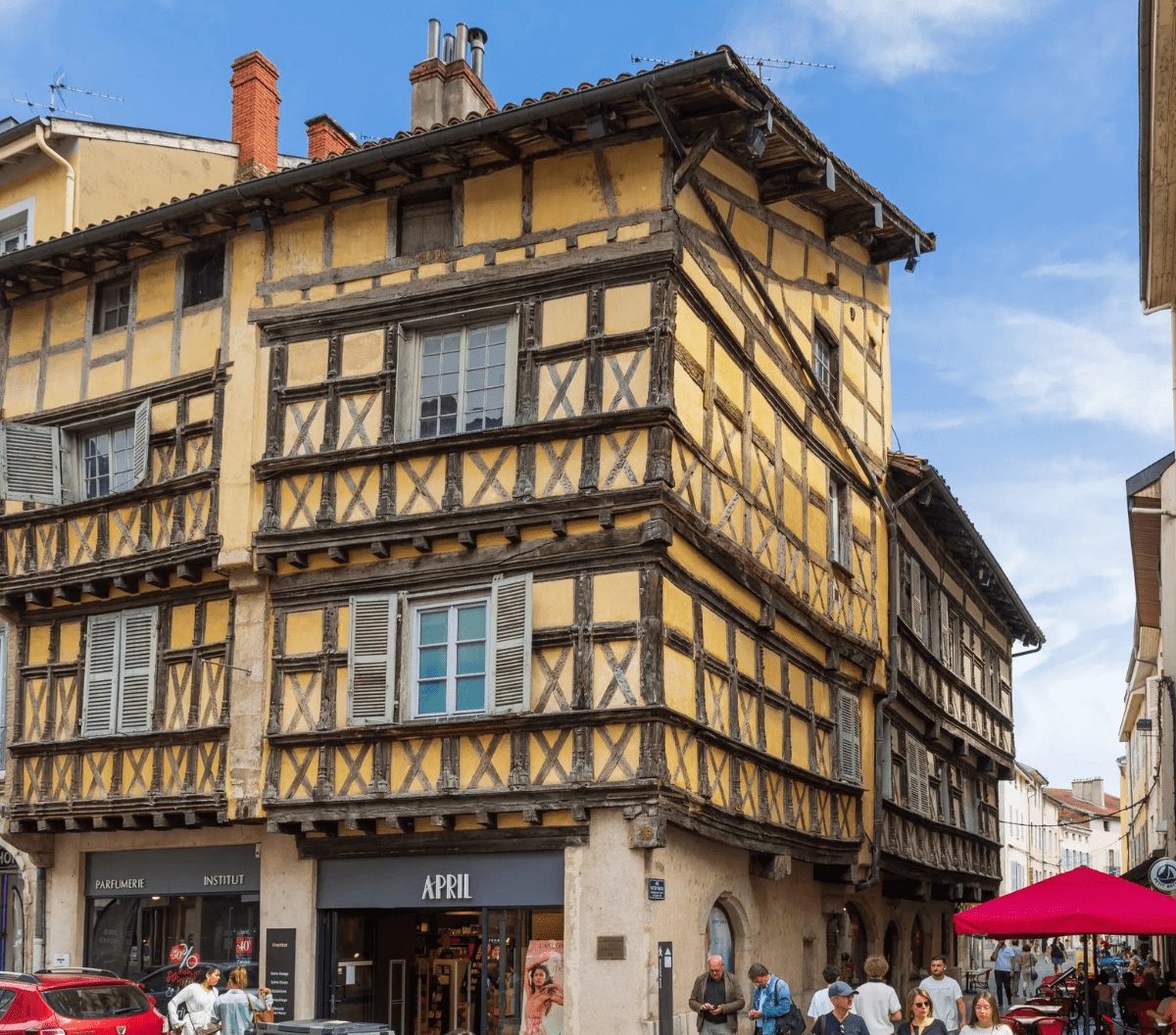 Un bâtiment historique jaune à colombages avec des poutres en bois dans une rue animée. Des gens passent devant des boutiques au niveau de la rue, et un parapluie rose vif ombrage des tables sur la droite. Ciel bleu avec des nuages au-dessus.