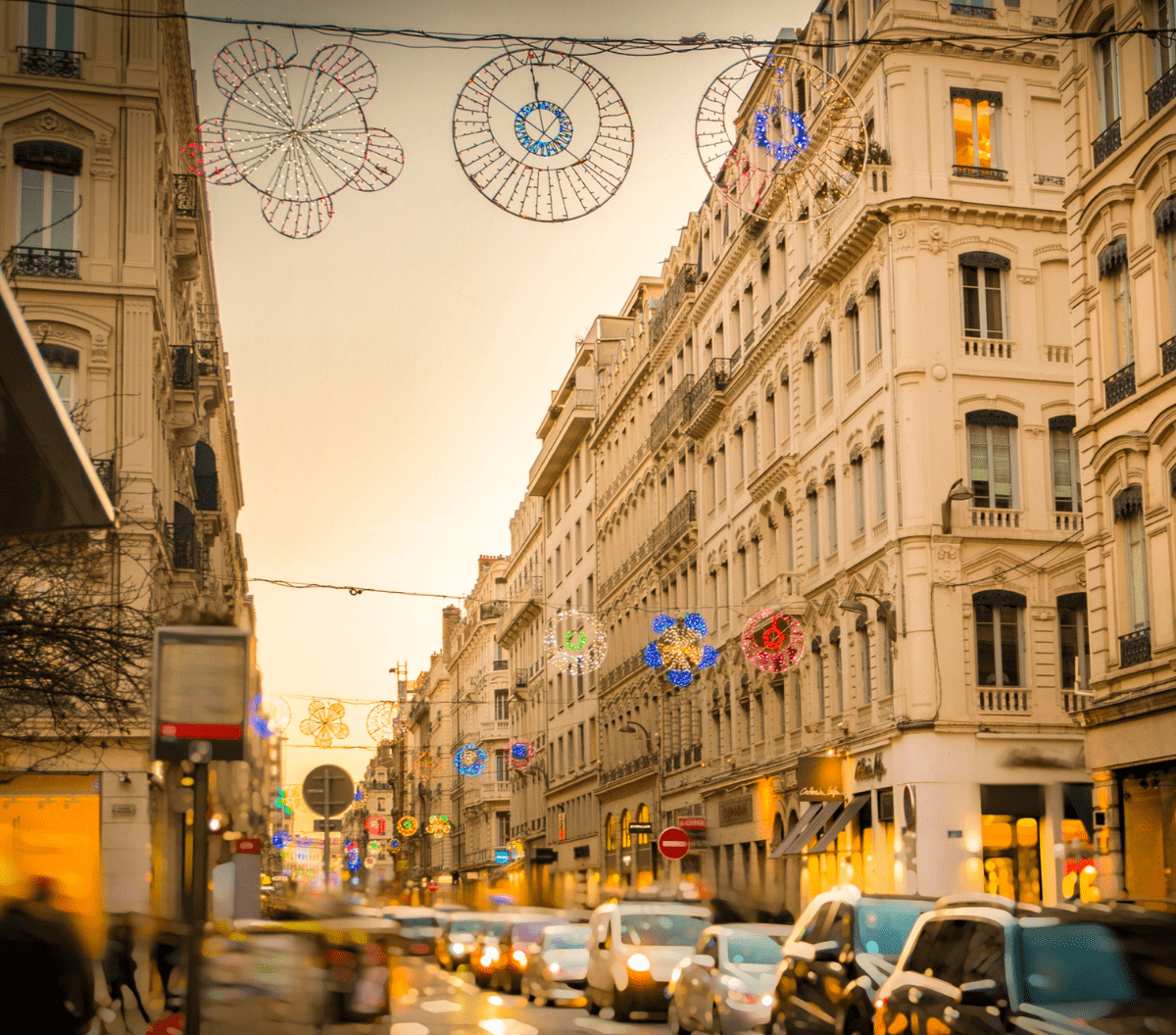 Une rue animée au coucher du soleil, avec des voitures et des gens, bordée de grands bâtiments élégants. Les lumières colorées des fêtes de fin d'année sont suspendues au-dessus, créant une atmosphère festive - une scène idéale pour mettre en valeur l'énergie vibrante d'investissement systématique à Lyon.